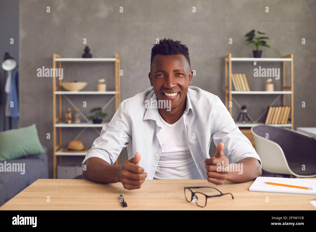 Felice amico giovane africano seduto al tavolo a casa ufficio e sorridendo alla macchina fotografica Foto Stock