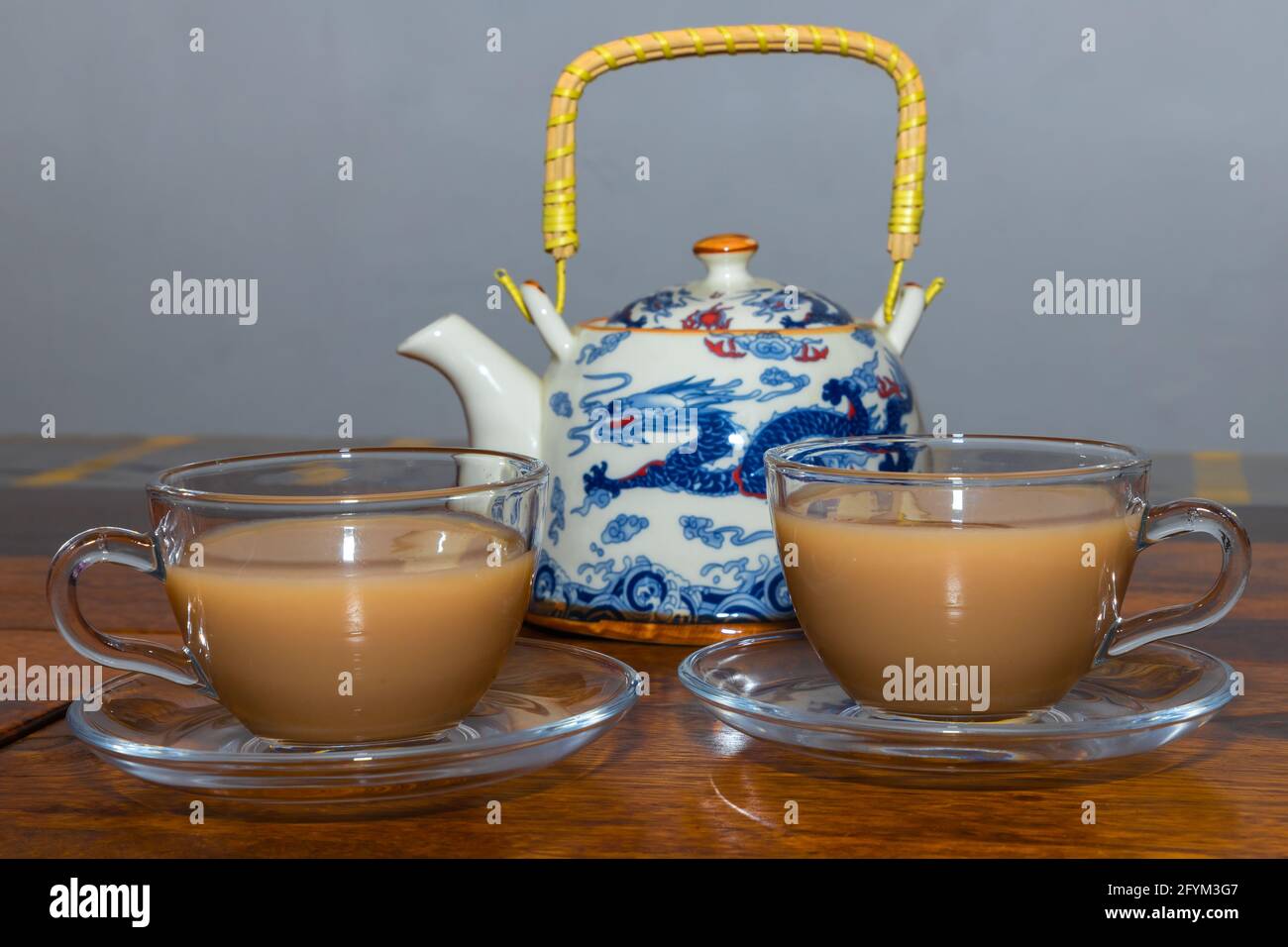 Tè fatto con latte servito in una tazza di vetro e piatto su un tavolo di legno da una teiera con manico in background Foto Stock