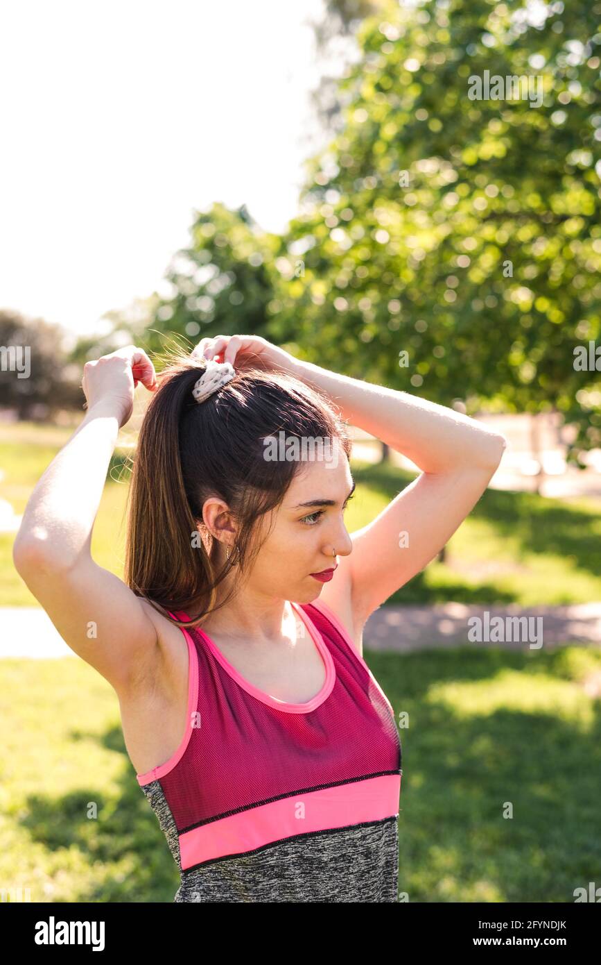 giovane caucasica che raccoglie i capelli con una coda di pony su un parco. Indossa ping e abiti sportivi verdi Foto Stock