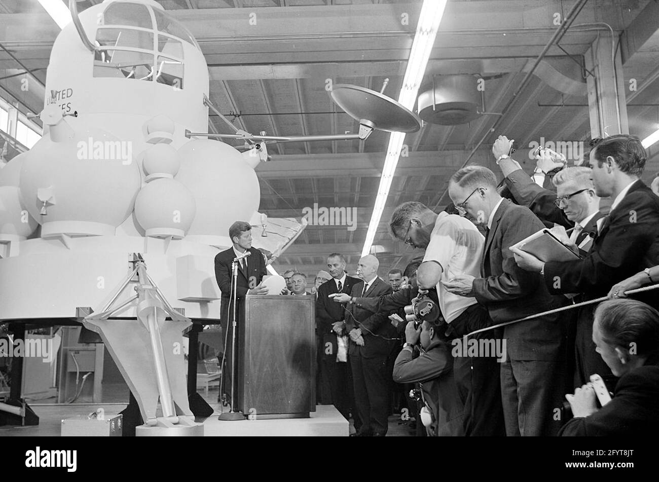 12 settembre 1962 il presidente John F. Kennedy (al leggio) trasmette le sue osservazioni, dopo un tour delle mostre di veicoli spaziali all'interno di un hangar presso il Rich Building del presidiato Spacecraft Center, Houston, Texas. Il presidente Kennedy possiede un modello in scala del modulo di comando Apollo, presentato a lui dal direttore del presidiato Spacecraft Center, il dottor Robert Gilruth; un mock-up del lander lunare (noto anche come 'il Bug') si trova a sinistra in background. In piedi: Direttore delle operazioni per il progetto Mercury, Dr. Walter C. Williams; amministratore associato della National Aeronautics and Space Administration Foto Stock