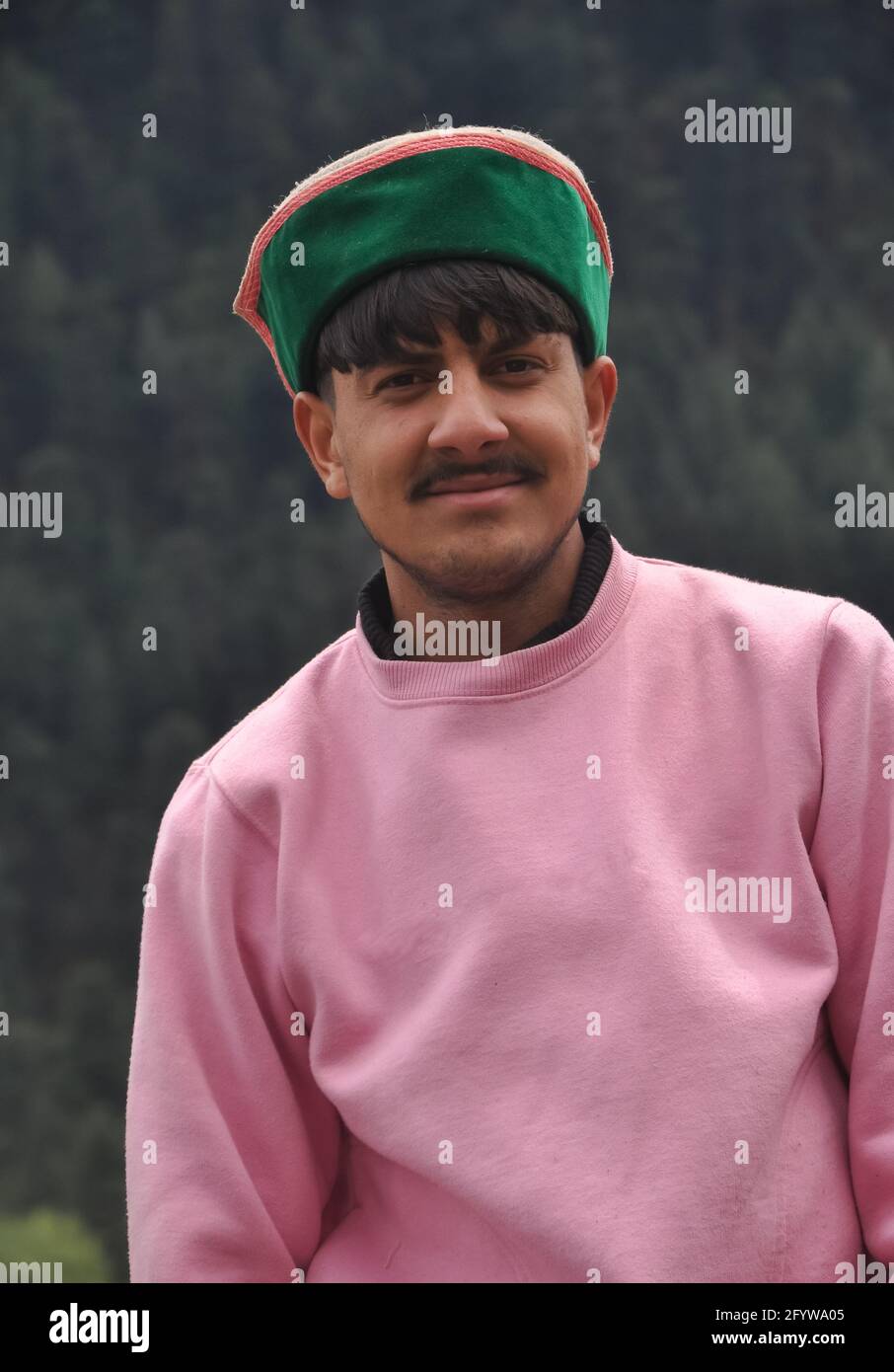 Ritratto di un bel giovane ragazzo che indossa felpa rosa e himachali topi (cappellino tradizionale Himachali), un bell'uomo indiano del nord in piedi all'aperto guardando la macchina fotografica Foto Stock