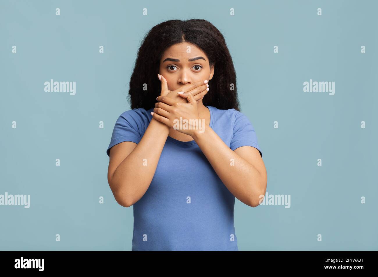 Ops. Donna afroamericana che ricopre la bocca con entrambe le mani e che sta fissando la fotocamera sullo sfondo blu dello studio. Foto Stock