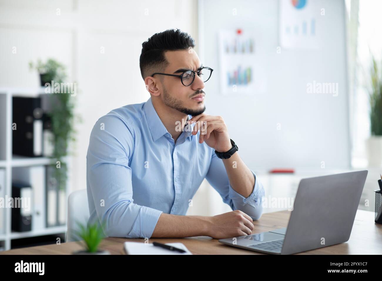 Un uomo d'affari arabo tormentato seduto alla sua scrivania, guardando lo schermo del portatile, risolvendo i problemi di lavoro in un ufficio moderno Foto Stock