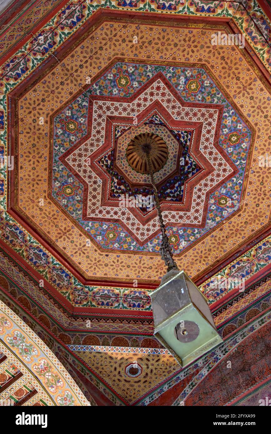 Lampada marocchina ornata è appesa a un soffitto dipinto in legno di cedro, noto anche come Zouak, a Palais Bahia, Marrakech, Marocco, Nord Africa. Foto Stock