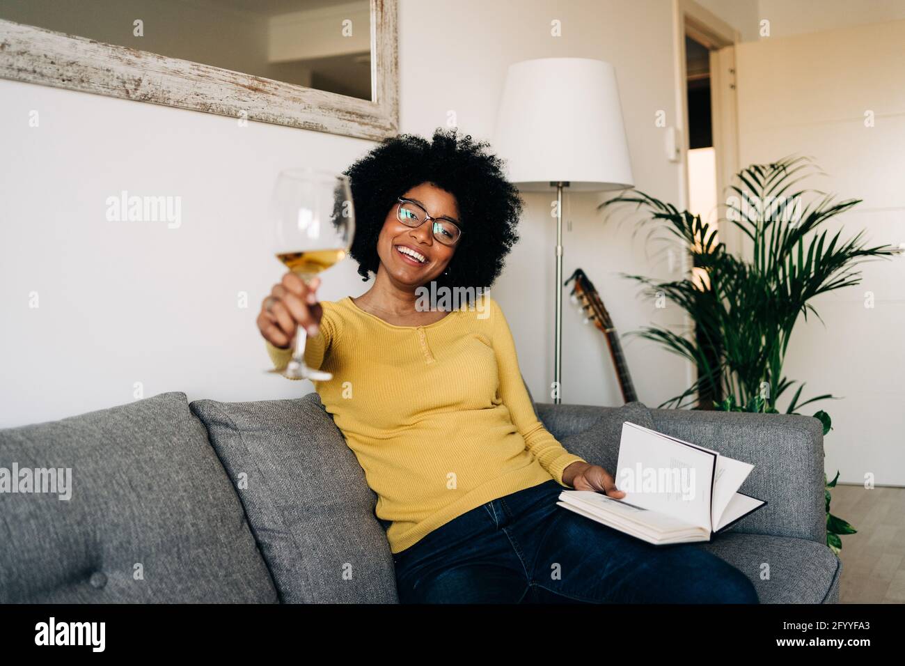 Felice donna afroamericana in giallino e maglione sul divano grigio morbido nel salotto Foto Stock