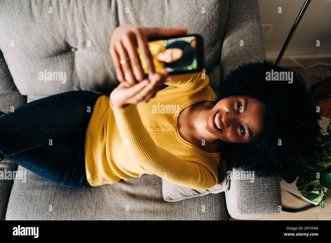 Vista dall'alto di una donna afro-americana felice che si autoritratti con il telefono cellulare mentre si sdraiano sul divano Foto Stock