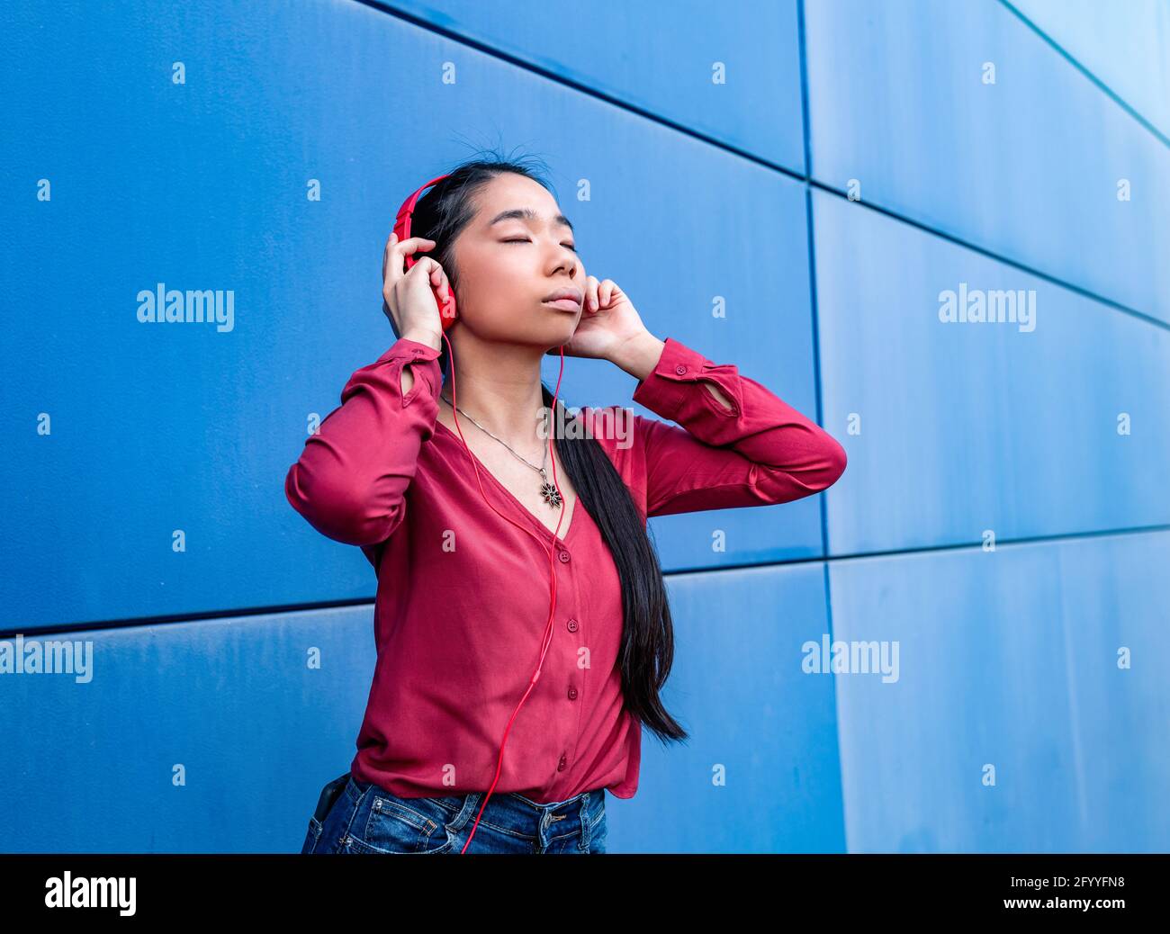Donna etnica con occhi chiusi che ascolta la musica in cuffia in piedi contro la parete blu Foto Stock