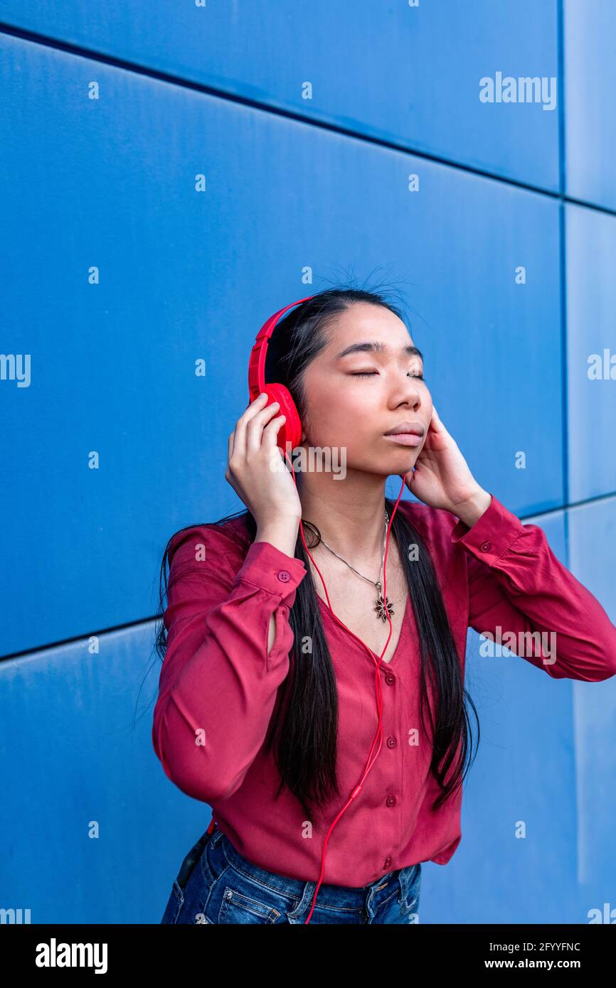 Donna etnica con occhi chiusi che ascolta la musica in cuffia in piedi contro la parete blu Foto Stock
