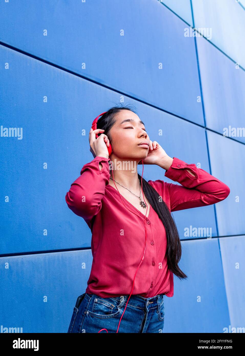 Donna etnica con occhi chiusi che ascolta la musica in cuffia in piedi contro la parete blu Foto Stock