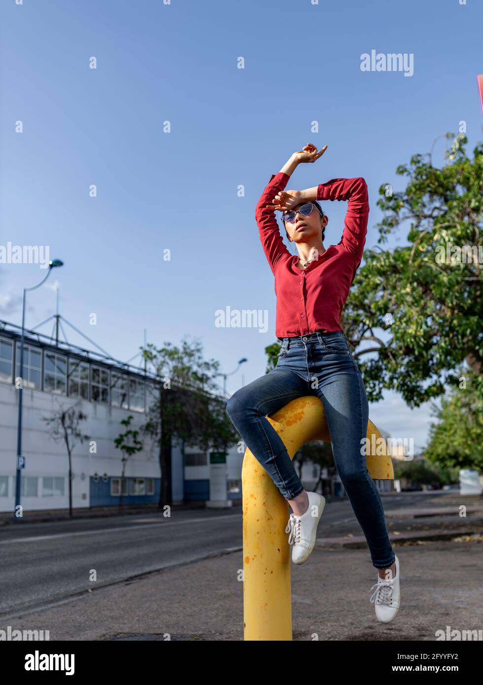Corpo sottile giovane donna asiatica in jeans e rosso blusa seduta con le braccia sollevate con grazia su tubo di metallo sopra strada sulla soleggiata giornata estiva Foto Stock