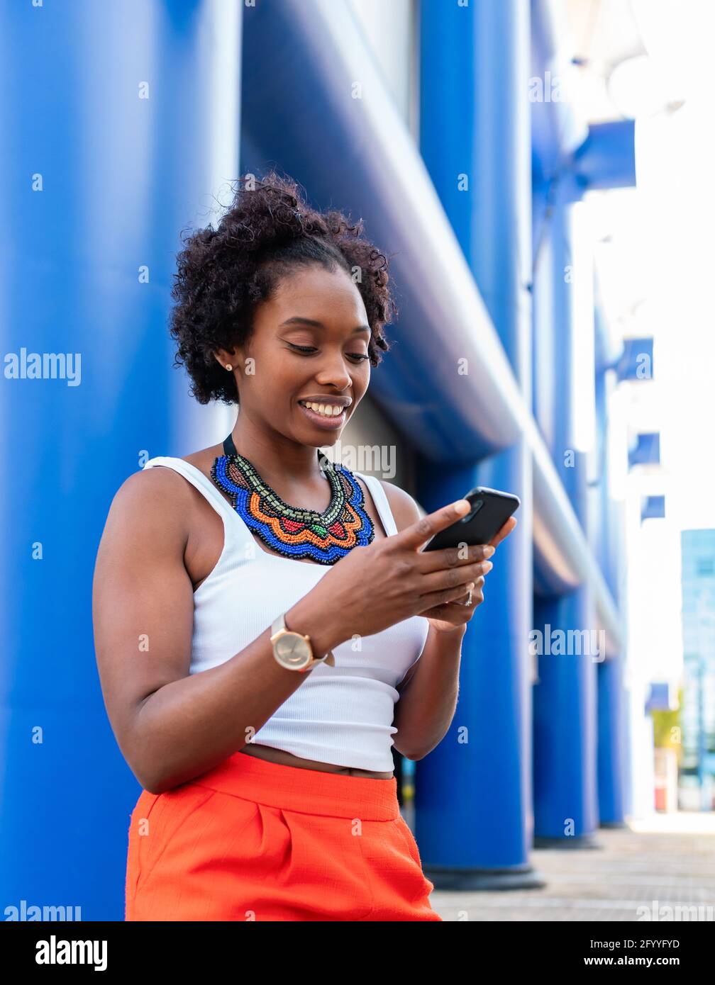 Giovane donna afro-americana allegra in estate shorts browsing mobile telefono e passeggiate in strada soleggiata Foto Stock