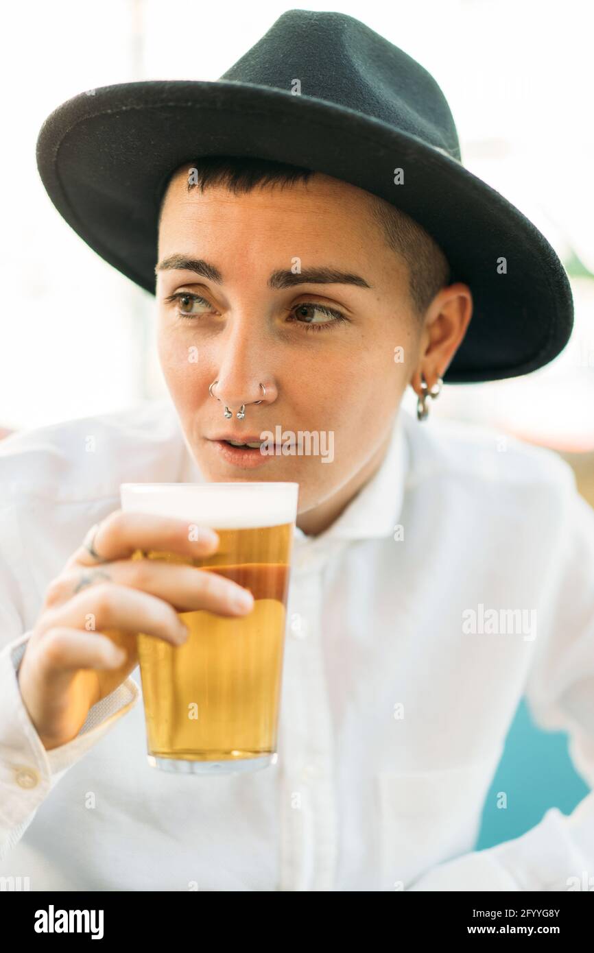 Tombino positivo con tatuaggi che indossano cappello e camicia bianca che beve birra dal bicchiere nel caffè Foto Stock