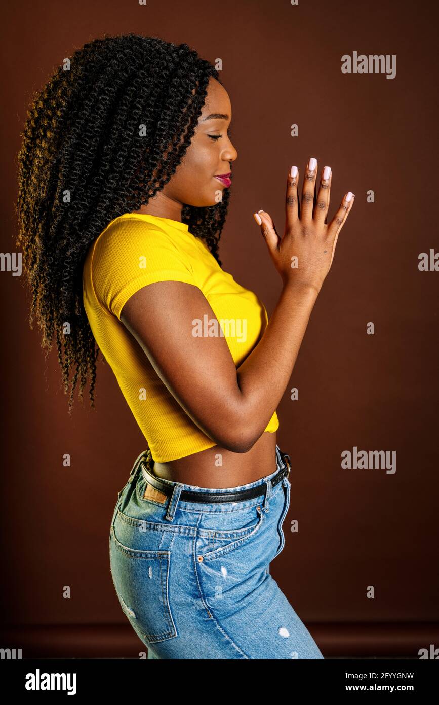 Vista laterale di una donna afro-americana sorridente in piedi con la preghiera mani e occhi chiusi in studio Foto Stock