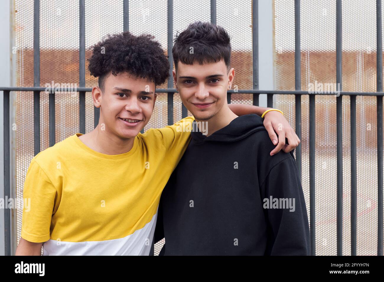 Sorridente uomo nero abbracciando l'amico teen mentre si sta in piedi vicino a. l'un l'altro contro recinzione metallica Foto Stock