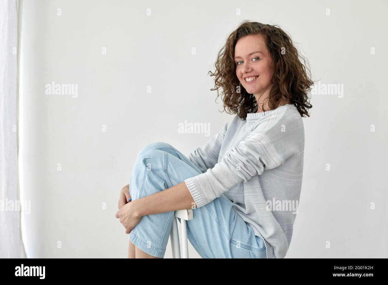 Vista laterale della giovane donna in abito alla moda seduta sedia di legno su sfondo bianco e guardando la macchina fotografica Foto Stock