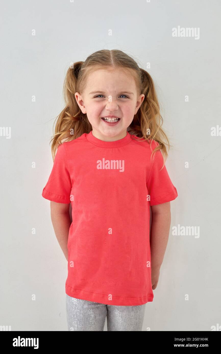Adorabile bambina allegra con coda di pony che indossa una maglietta rosa in piedi su sfondo bianco e guardando la fotocamera con un sorriso Foto Stock