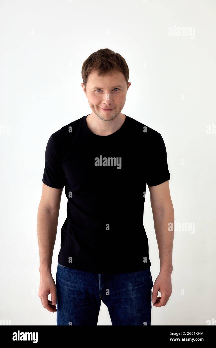 Felice giovane maschio in jeans e maglietta nera in piedi su sfondo bianco e guardando la fotocamera Foto Stock