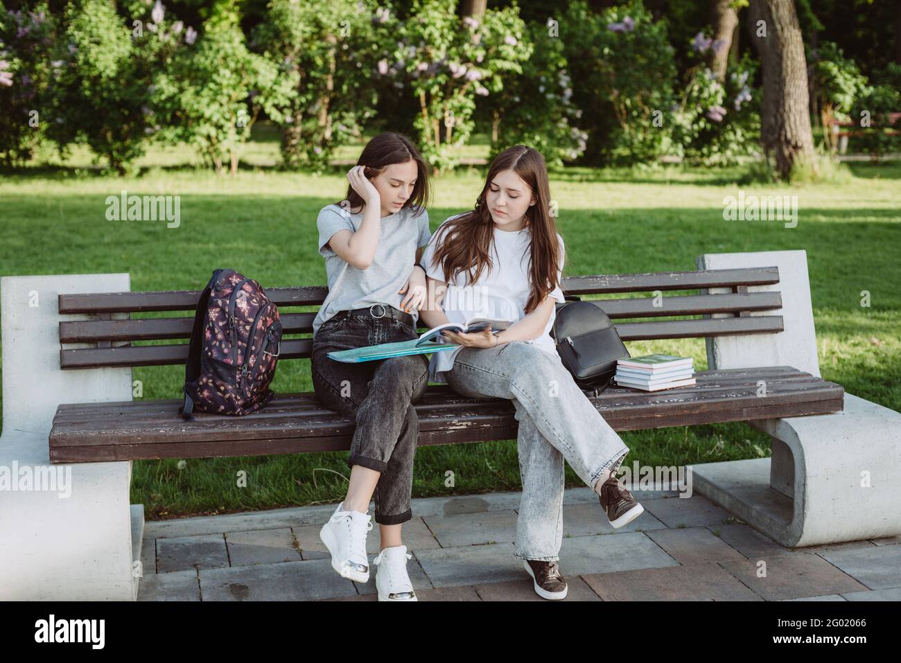 Due ragazze studentesche stanno guardando un libro aperto su una panchina nel parco. Formazione a distanza, preparazione agli esami. Messa a fuoco selettiva morbida. Foto Stock