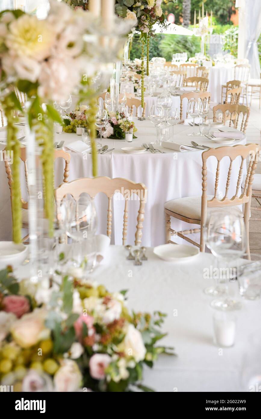 Luogo di nozze all'aperto in un giardino con arredamento elegante e posti su tavoli bianchi decorati con centrotavola floreale, bicchieri e stoviglie Foto Stock