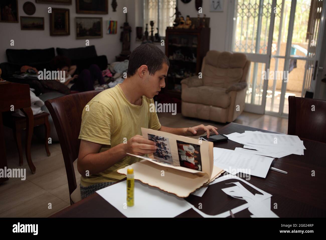 il giovane di 22 anni è un messaggio di testo con il suo telefono cellulare, capovolge le pagine di un album di famiglia mentre si siede e studia nel soggiorno a casa. Foto Stock