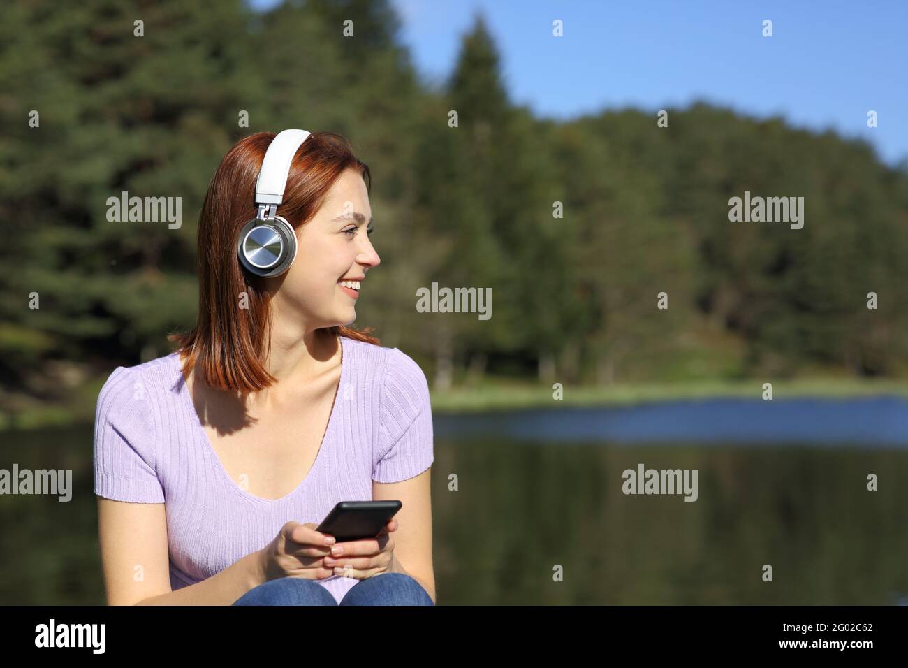 Donna felice che ascolta la musica con il telefono e le cuffie wireless guardando a lato in un lago Foto Stock