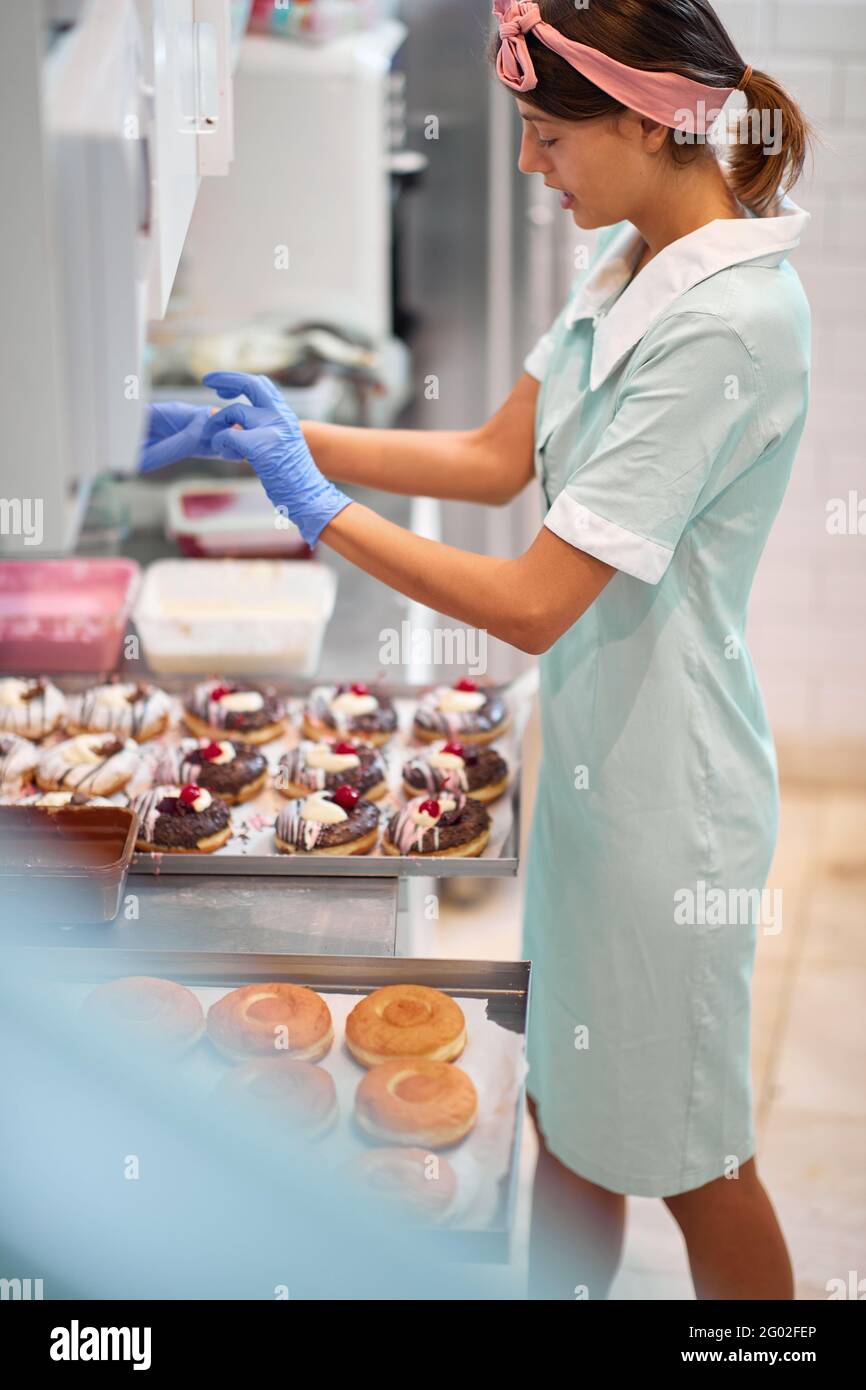 Una giovane piccola donna di affari è godendo nella preparazione delle ciambelle squisite fatte a mano in un'atmosfera rilassata in un laboratorio. Pasticceria, dessert, dolce, Foto Stock
