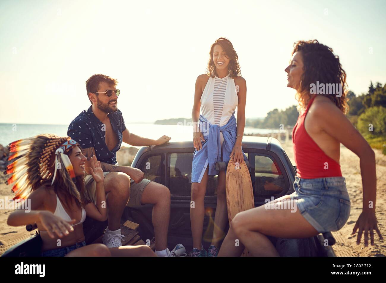 Un gruppo di bei modelli è seduto sul retro di un'auto vicino al mare e chiacchierando in una bella giornata di sole. Estate, mare, vacanza, amicizia Foto Stock