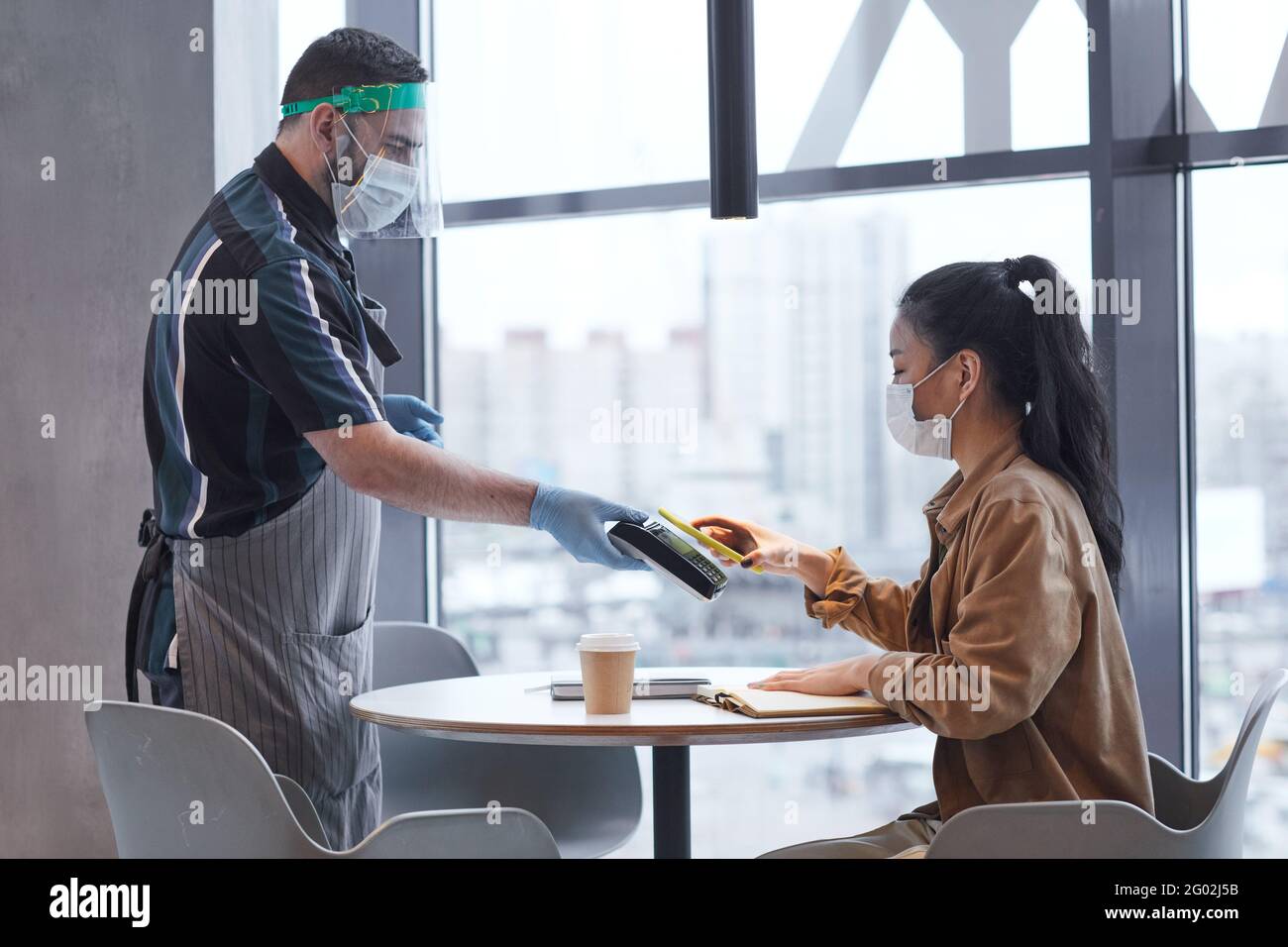 Vista laterale verticale del cameriere che indossa visiera e guanti mentre serviva la cliente femminile in un bar con misure di sicurezza covide Foto Stock