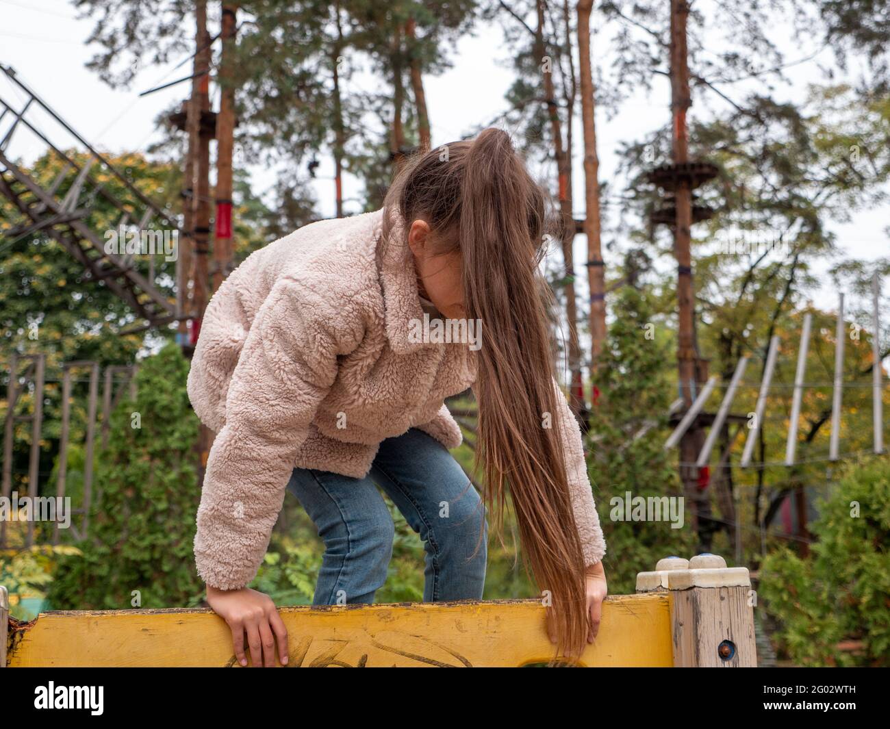 Felice ragazza sorridente adolescente in un cappotto di pelliccia sintetica e con lungo ponytail che sale l'arrampicatore giallo al parco giochi per bambini. Parco di corda con alberi di pino Foto Stock