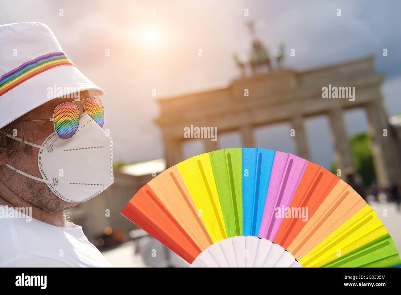 Uomo LGBT caucasico maturo a Berlino con ventola arcobaleno e nastro su cappello estivo bianco. Rainbow, simbolo dell'orgoglio gay LGBTQIA, la diversità. L'attivista maschile è Foto Stock