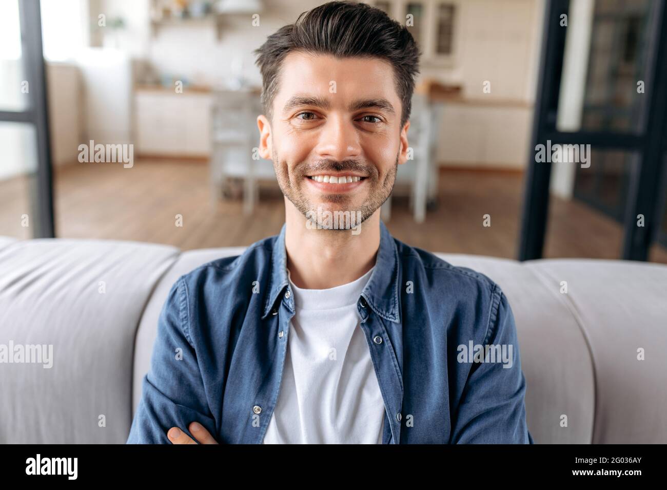 Primo piano ritratto di un bel giovane adulto caucasico, seduto a casa con le braccia incrociate sul divano, vestito in una casual elegante camicia in denim, guardando la macchina fotografica e sorridendo amichevole Foto Stock