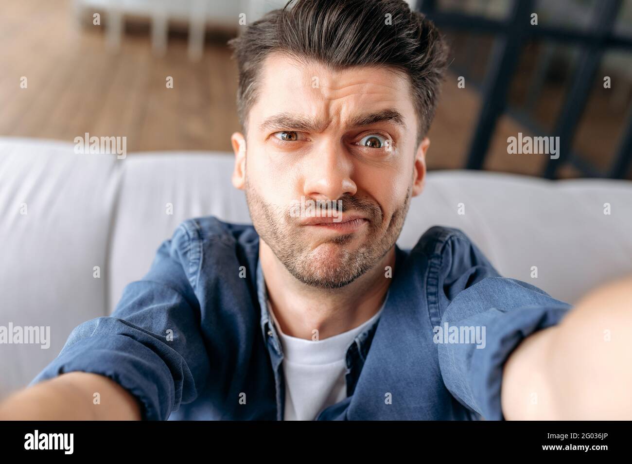 Ritratto di un giovane adulto perplesso uomo caucasico in abiti casual seduto sul divano, prendendo un selfie, grimacing durante le riprese, utilizza uno smartphone, imbrogliare intorno, guardando la webcam frontale Foto Stock