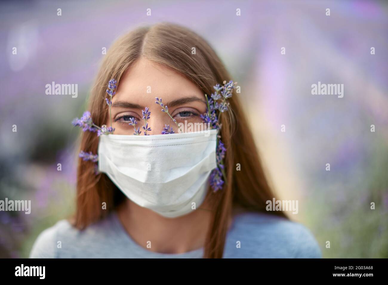 Ritratto di giovane donna attraente che indossa maschera protettiva con fiori all'interno guardando la macchina fotografica. Bella modello femminile in posa in campo lavanda, belle macchie sullo sfondo. Concetto pandemico. Foto Stock