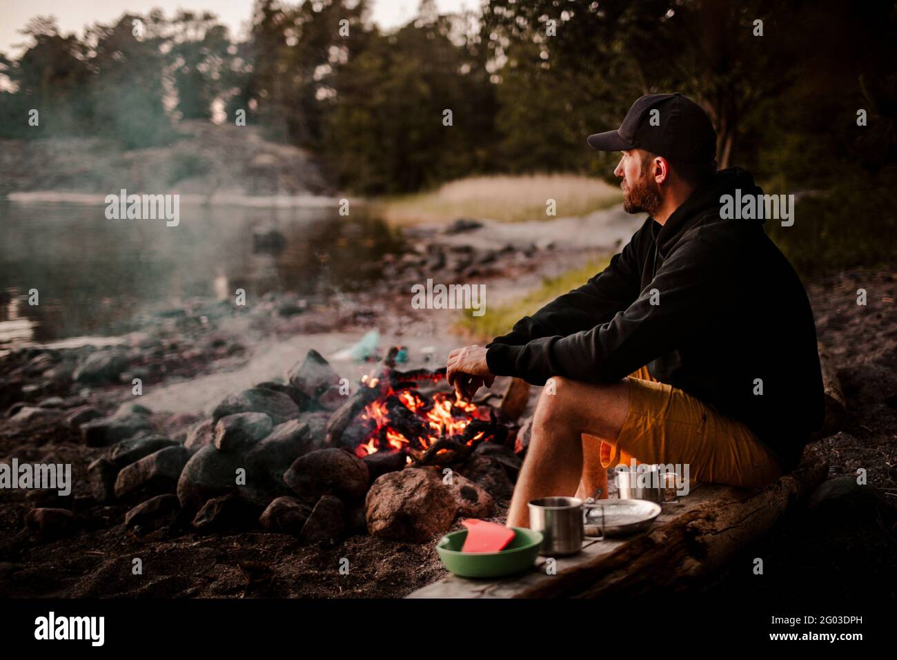 Vista laterale di un uomo maturo seduto accanto al fuoco mentre si guarda lontano durante il tramonto Foto Stock