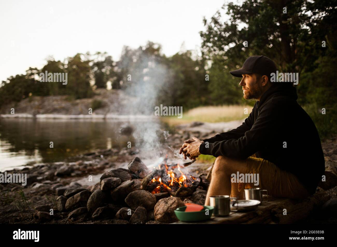 Vista laterale di un uomo attento che guarda lontano mentre si siede accanto al fuoco sul lungolago Foto Stock