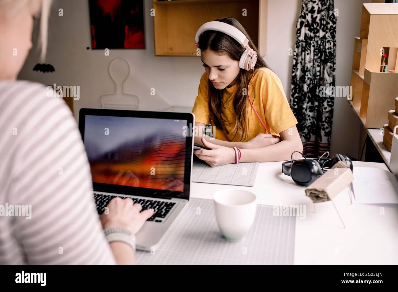 Ragazza che indossa le cuffie e-learning attraverso il tablet digitale mentre si siede con una donna d'affari che lavora su un computer portatile alla scrivania Foto Stock