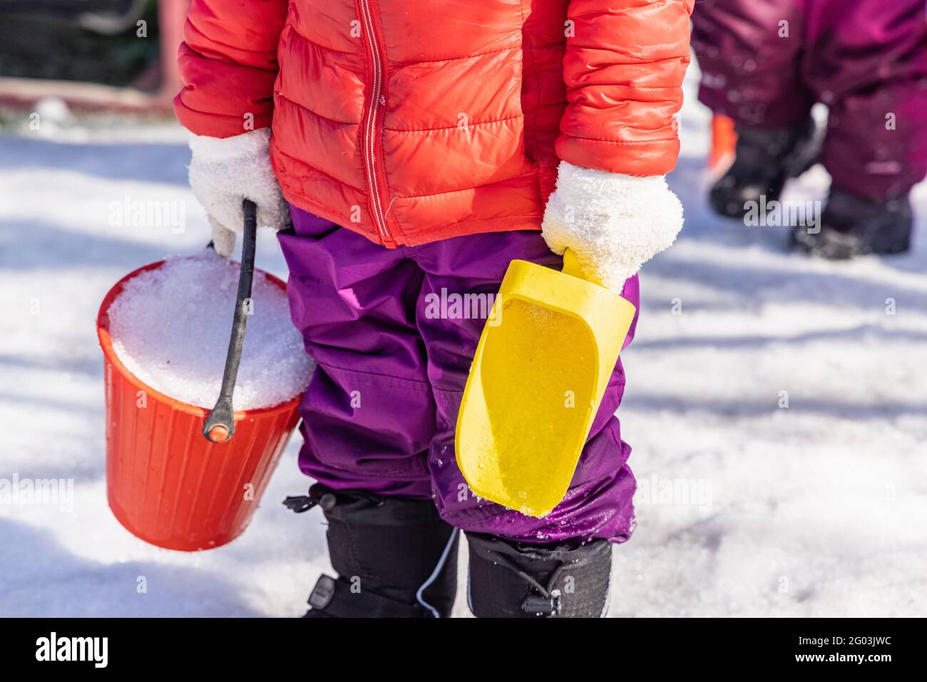 Primo piano di un bambino irriconoscibile giovane asilo in abiti invernali e giacca che gioca con un secchio di plastica rosso e una paletta gialla. Foto Stock
