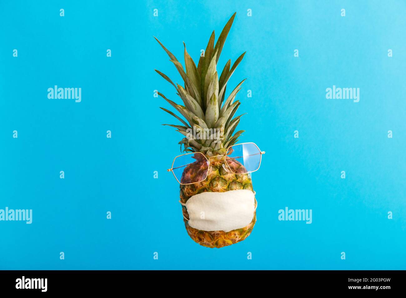 Ananas divertente faccia in occhiali da sole maschera medica di stile su sfondo blu estate. Concetto di viaggio ananas in maschera protettiva in coronavirus Foto Stock