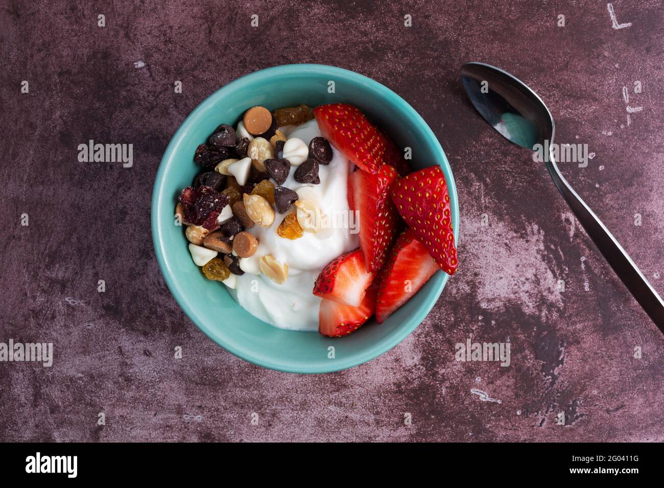 Vista dall'alto di una ciotola con yogurt greco puro e un mix di caramelle e fragole su un piano di un marronone con un cucchiaio a lato. Foto Stock