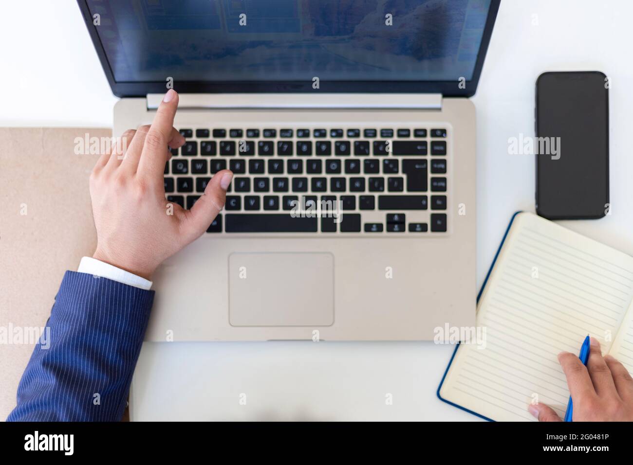 Posa piatta, mano di uomo d'affari che scrive su notebook con computer portatile e cellulare, raccoglitore e penna sulla scrivania in ufficio. Colpo di testa Foto Stock