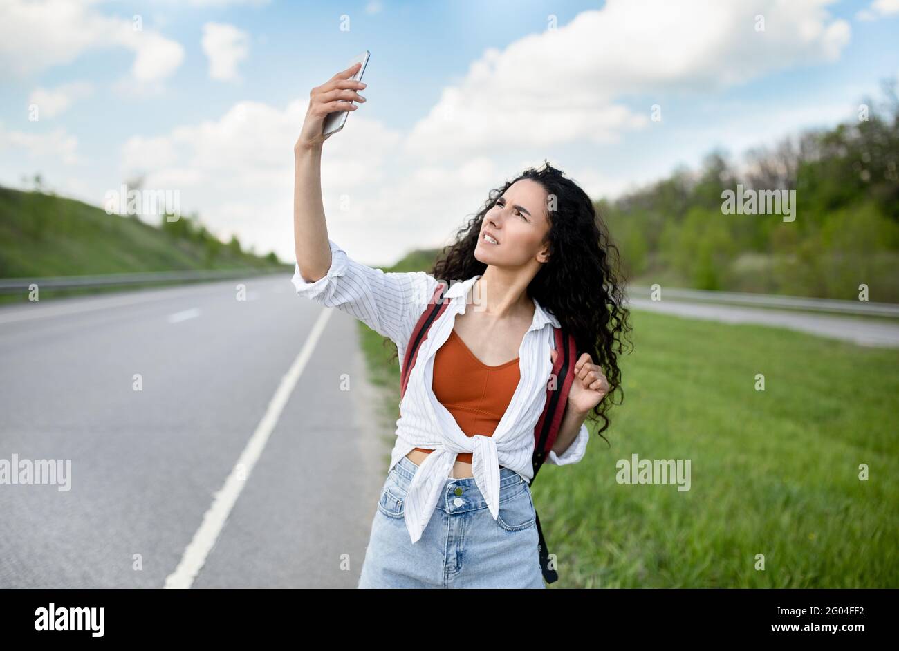 Turbata giovane donna che cammina lungo la strada, alzando la mano con smartphone, alla ricerca di segnale, senza connessione, all'aperto Foto Stock