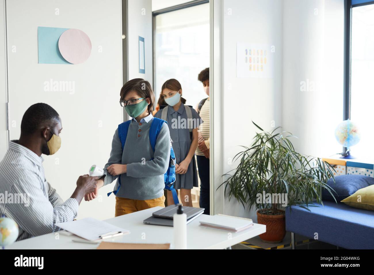 Gruppo di bambini che indossano maschere che entrano in classe con l'insegnante che controlla la temperatura con il termometro a infrarossi, sicurezza covid Foto Stock