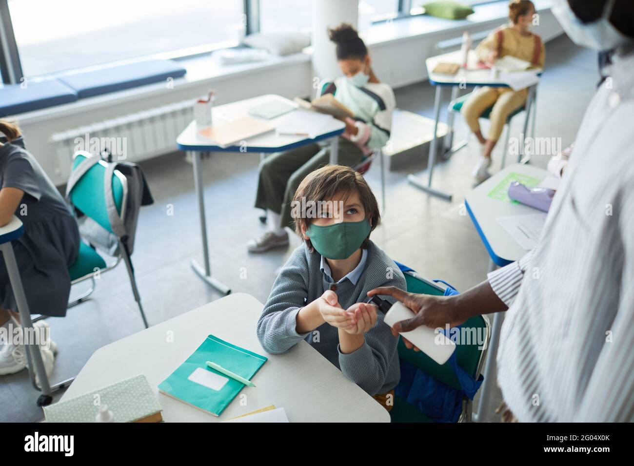 Ritratto ad alto angolo del ragazzo che indossa una maschera e mani igienizzanti in classe scolastica, spazio copia Foto Stock