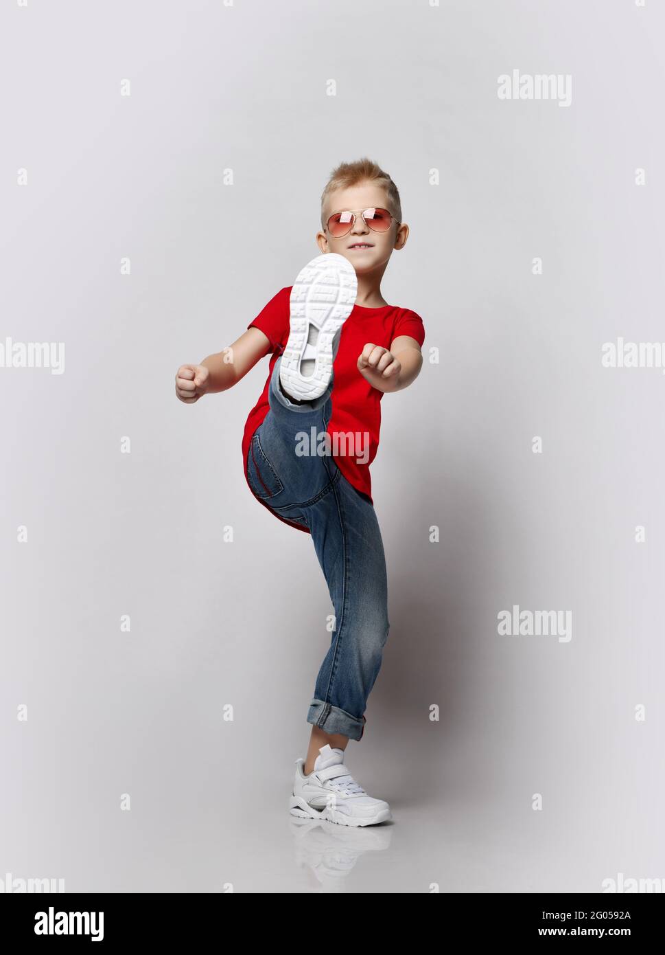 Elegante ragazzo allegro in t-shirt rossa, jeans e occhiali da sole che fanno calcio con gamba guardando la fotocamera Foto Stock