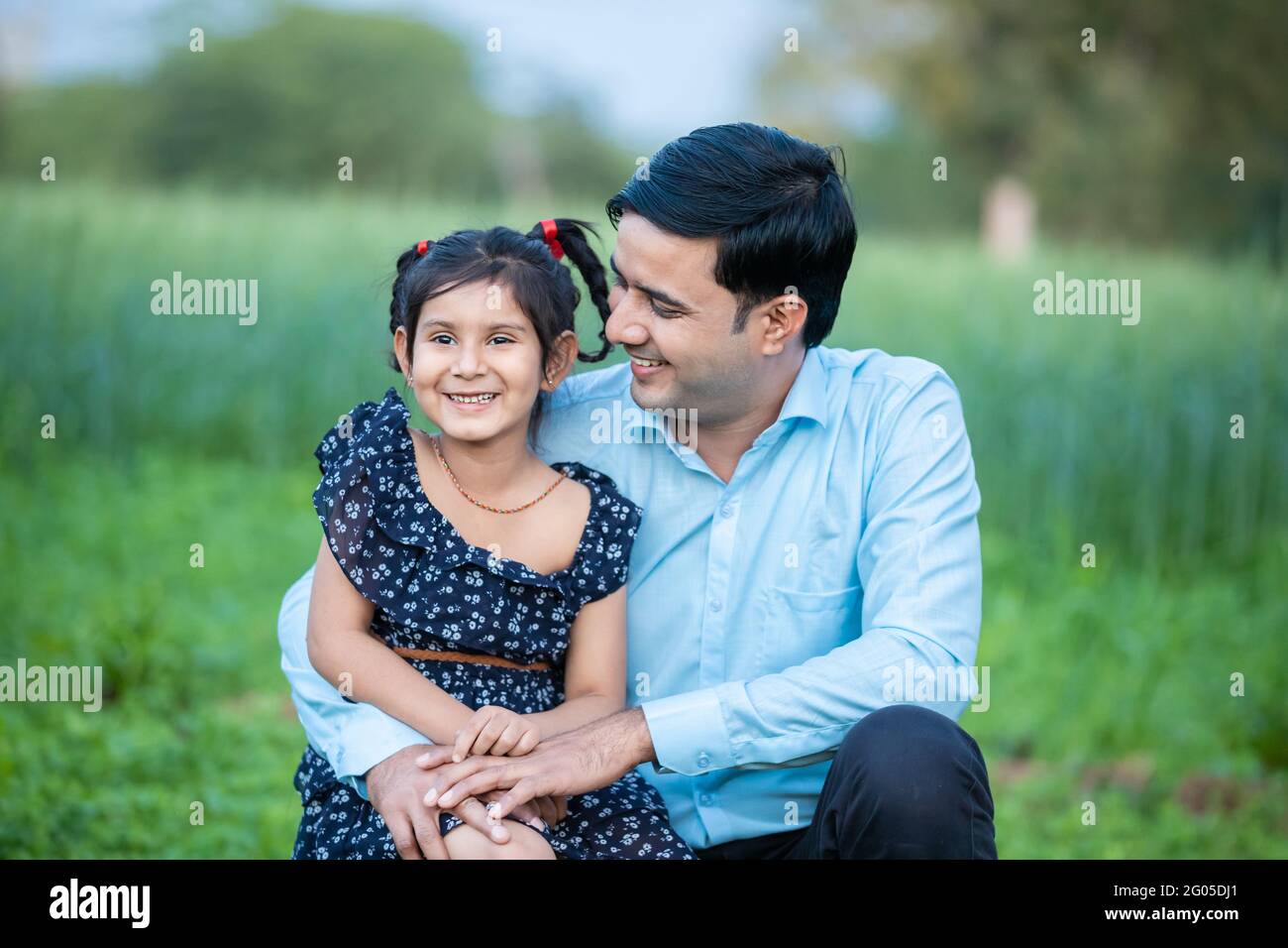 Felice sorridendo giovane indiano più lontano abbracciando la sua piccola figlia carina seduta all'aperto a verde agricoltura campo sfondo, legame, amore, relazione Foto Stock