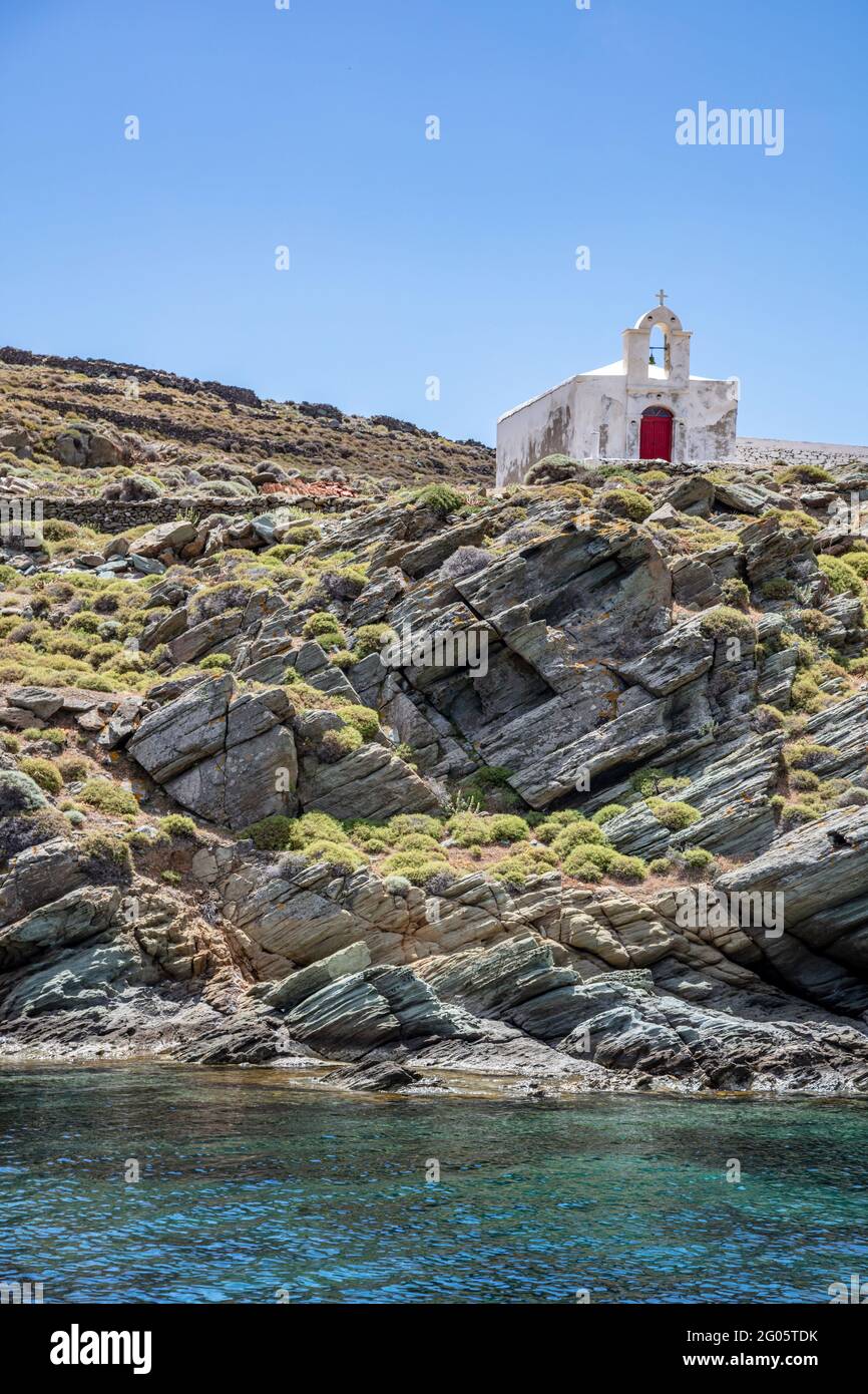 Piccola chiesa nell'isola di Kythnos, Cicladi, Grecia. Cappella ortodossa dipinta di bianco con pareti sbiadite con campanile e porta rossa su una scogliera rocciosa su blu chiaro s. Foto Stock