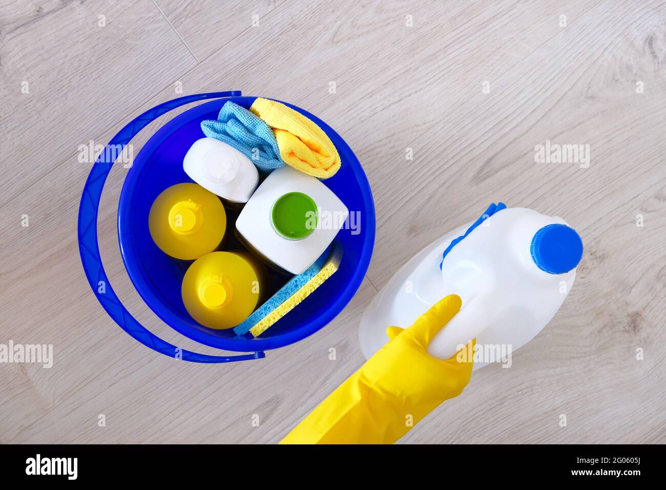 Detersivi in bottiglie di plastica, spugne, spazzola in secchio blu per uso domestico. Mano nel guanto di gomma giallo. Concetto di servizio. Vista dall'alto. Disposizione piatta. Foto Stock