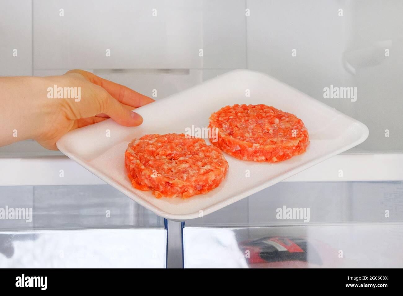 Il Burger di pollo crudo in contenitore è su ripiano in frigorifero aperto. La mano femminile tira fuori dal frigorifero un ingrediente per cucinare piatti di carne. Foto Stock