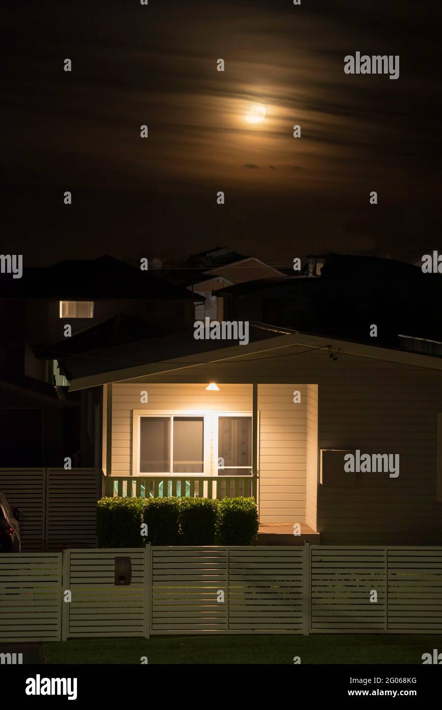 Una luna frizzante dietro le nuvole che si innalza sopra i tubi flessibili con una casa in primo piano con la sua luce anteriore accesa in Australia Foto Stock