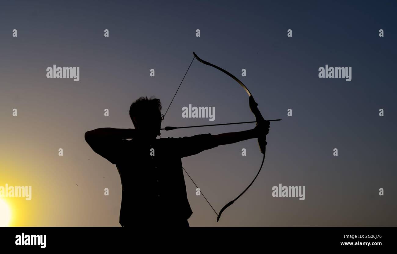 un uomo dalla silhouette che tiene un arco e una freccia davanti al tramonto. Foto Stock
